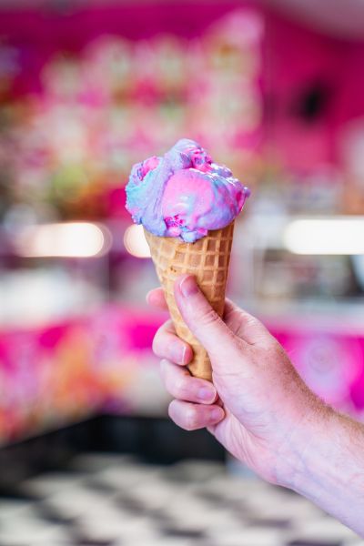 A hand holds an ice cream cone with a scoop of pink and blue ice cream. The background is a colorful ice cream shop.