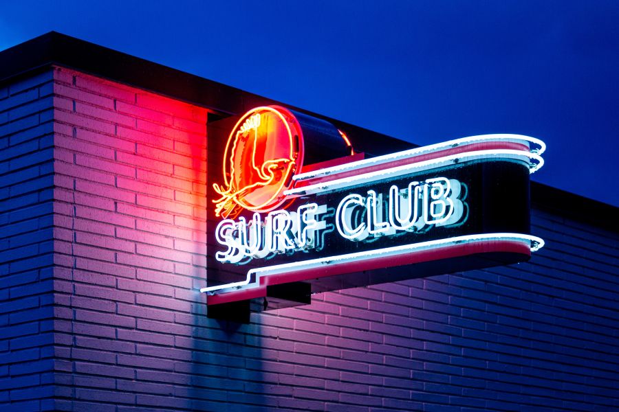 The neon store sign for Surf Club shines bright at dusk.