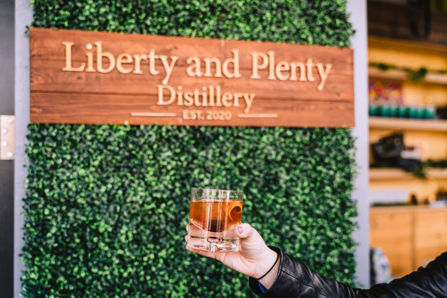 A hand holds up a cocktail in front of the Liberty and Plenty Distillery Sign.