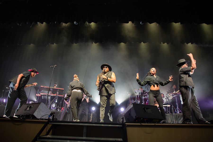 A band performs onstage. Five musicians all on different instruments.