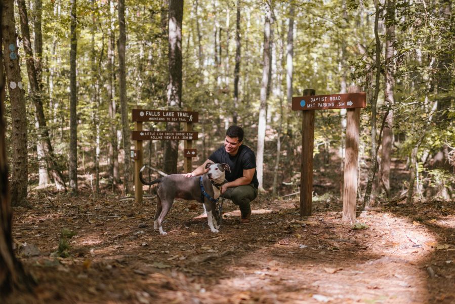 A man kneels with a dog in front of trail signs and trees at Falls Lake.