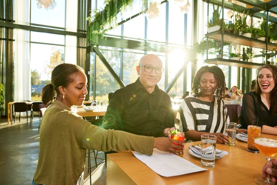 A group of professionally dressed people sit at a bar in a beautiful restaurant filled with plants and sunlight.