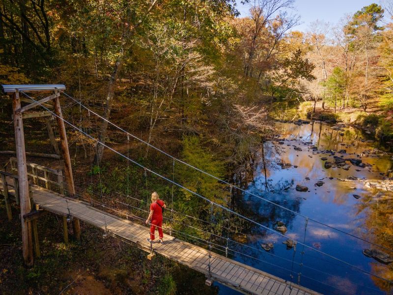 A woman in a red jumpsuit crosses a suspension bridge over the Eno River in the fall.