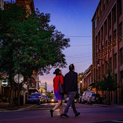 A couple walks the streets of Durham at dusk, framed by string lights hanging between two brick buildings.