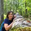 Writer Vanessa Infanzon poses for the camera in a forest, leaning on a large tree trunk